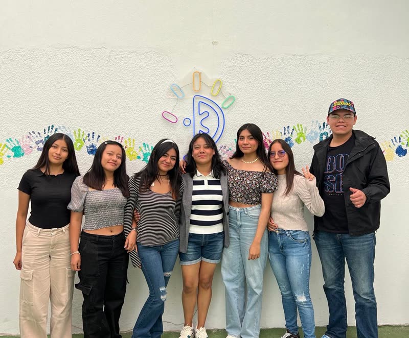 Alumnas y alumnos de CMUKA frente al muro con el logo de neón